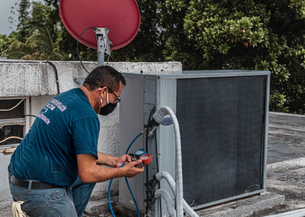 Technician repairing an air conditioner unit outdoors, wearing a facemask and using a manifold gauge